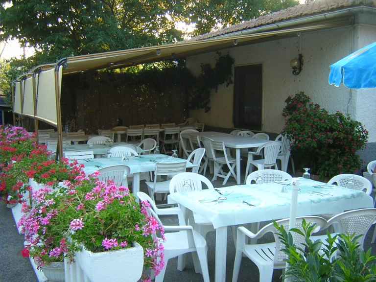 Outdoor patio dining area with white plastic tables and chairs under a canopy awning, surrounded by potted flowers