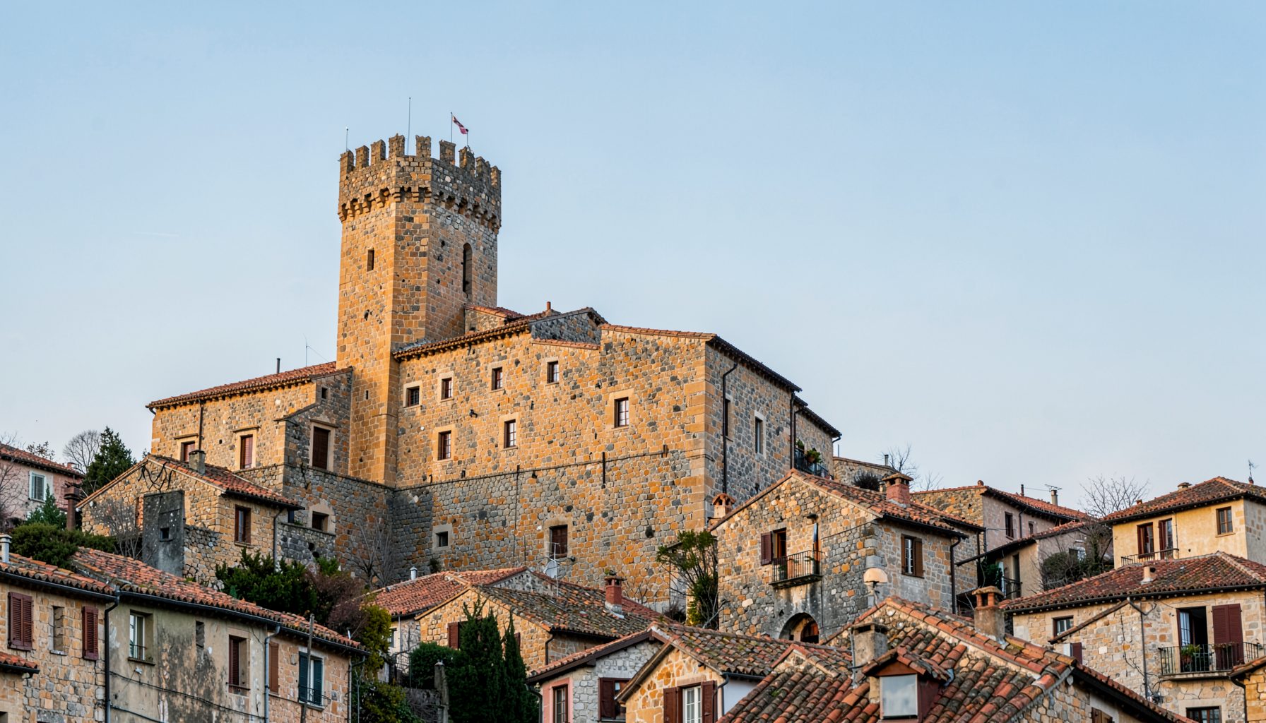 A fortified stone palace with a crenellated tower rising above the rooftops of a medieval hilltop village, warm ochre and grey stone against a soft blue sky