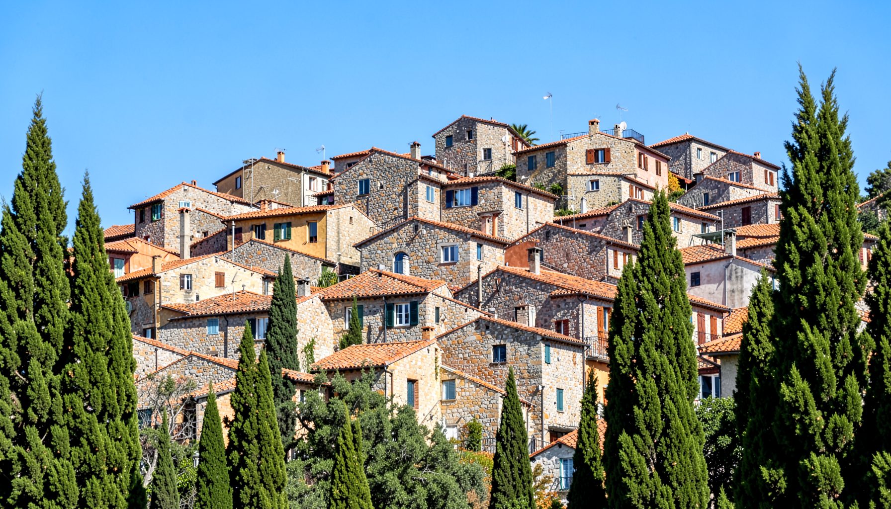 Dense cluster of stone buildings with terracotta roofs climbing a hillside, warm ochre and grey tones, framed by green cypress trees under a bright blue sky